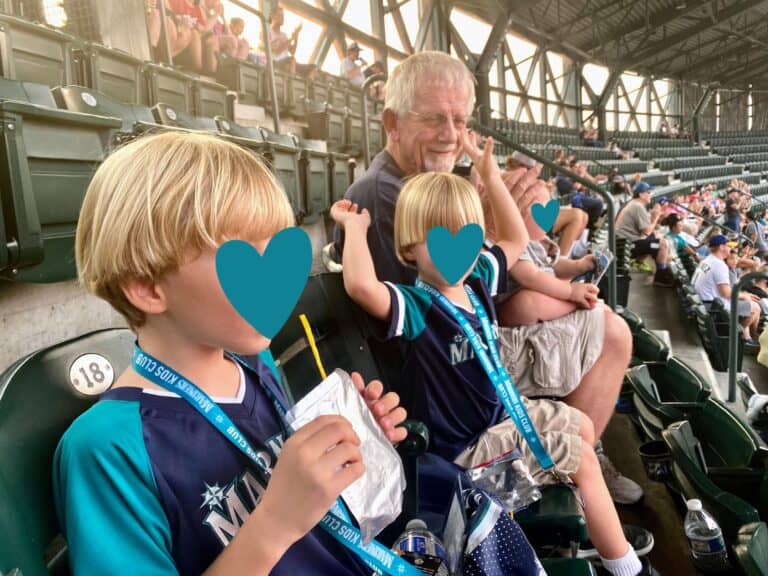 Grandfather smiles while holding baby and looking at his grandsons in the stands of Seattle Mariners baseball stadium.