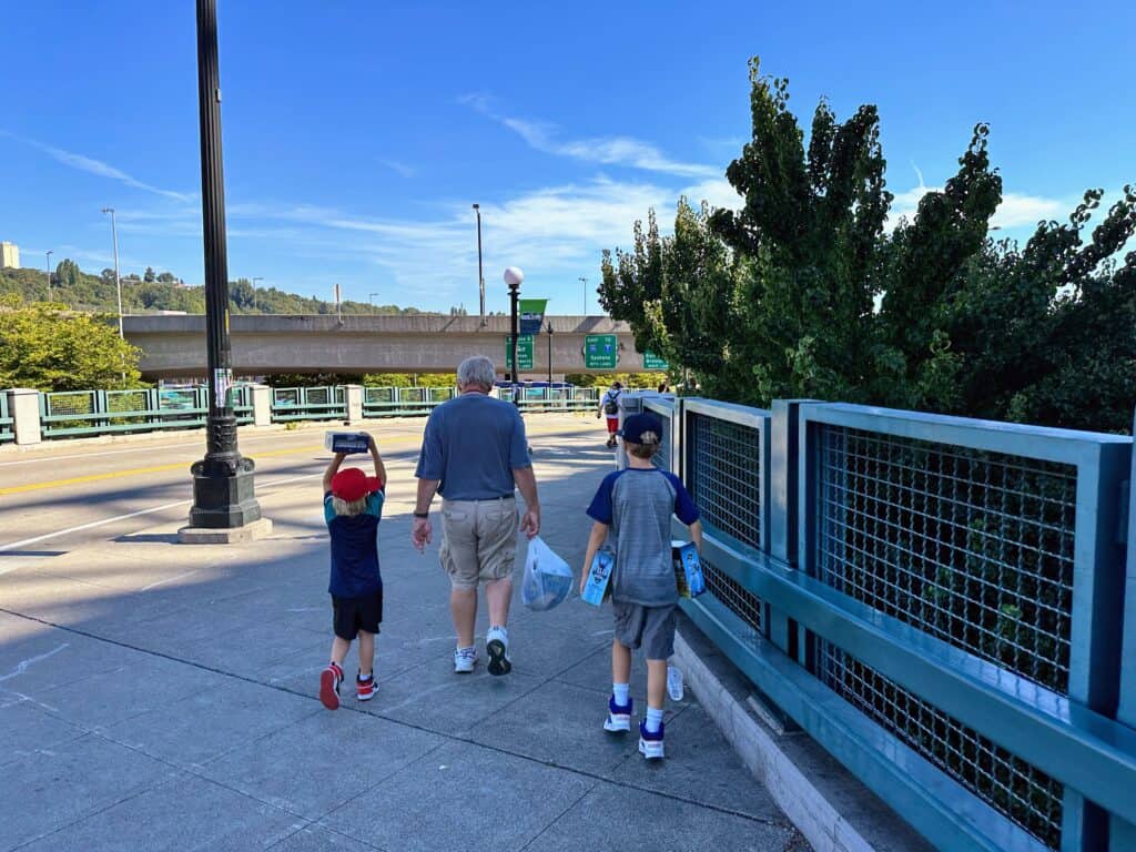 Grandfather walks on sidewalk with one grandson walking on each side of him. They are outside the Seattle Mariners baseball stadium.