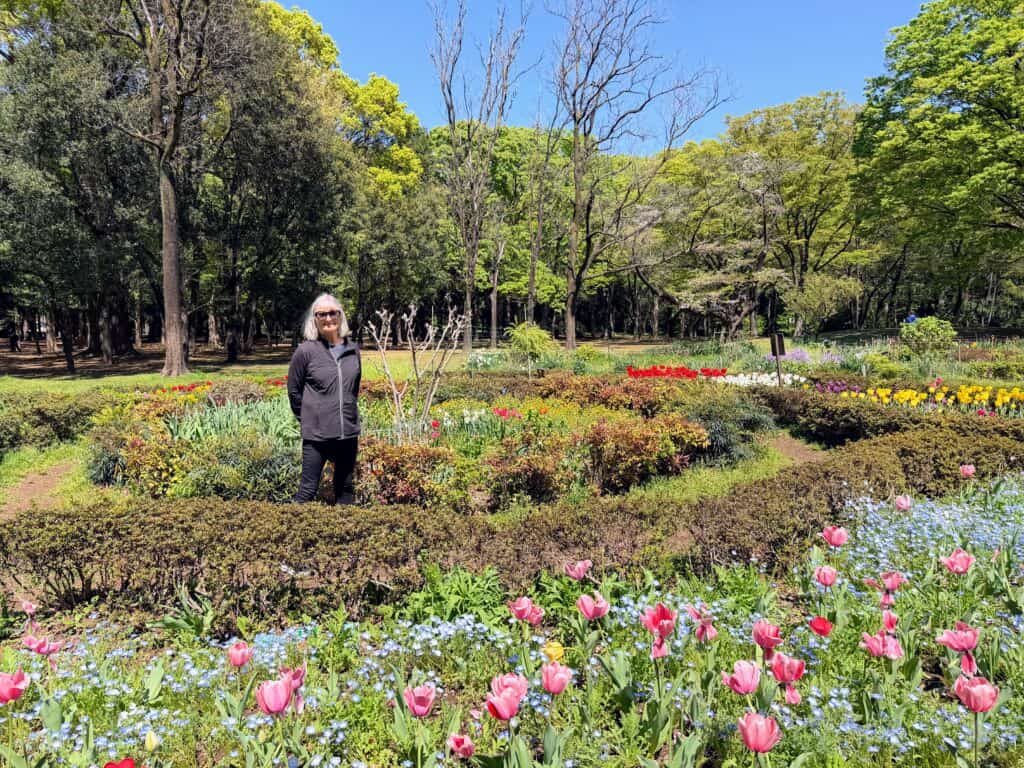 Woman stands in the middle of a colorful flower garden in a park in Tokyo, Japan. Behind her are larger trees.