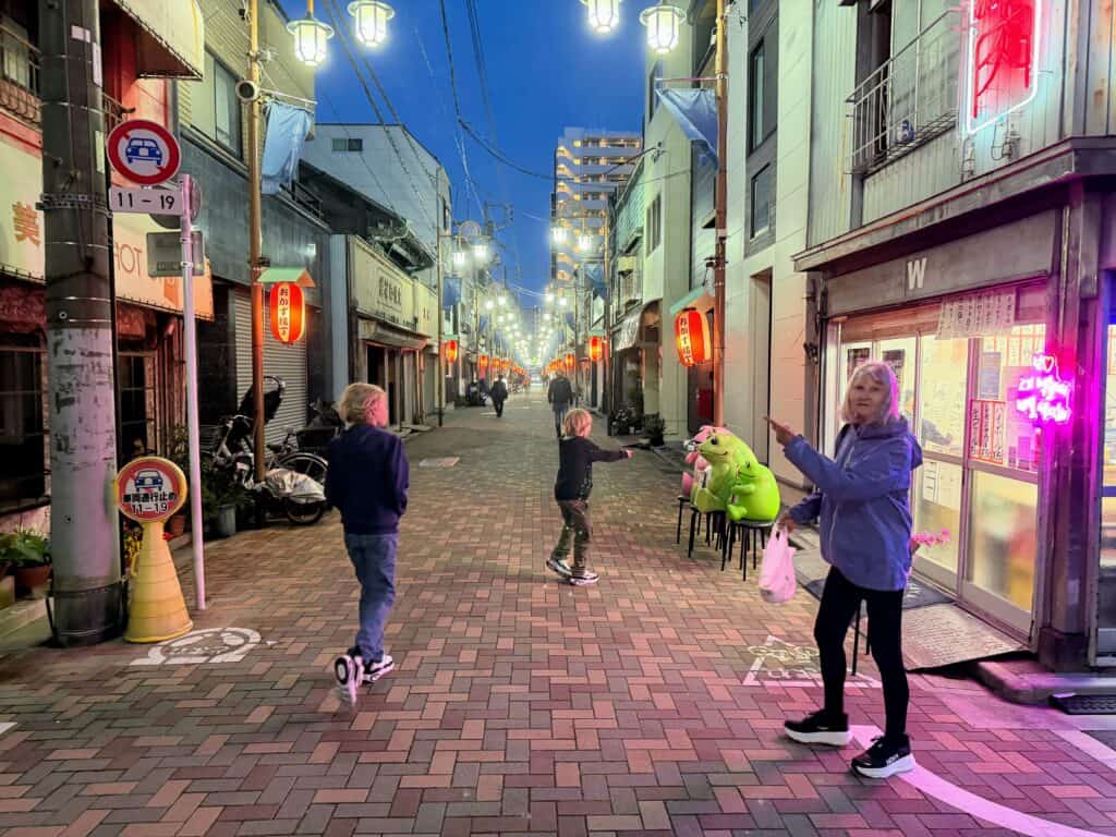 Woman points to the left and boys walk to her right down a street. It's dusk in Tokyo. The boys are wearing sweatshirts and looking at large plush frogs sitting in chairs in front of a shop.