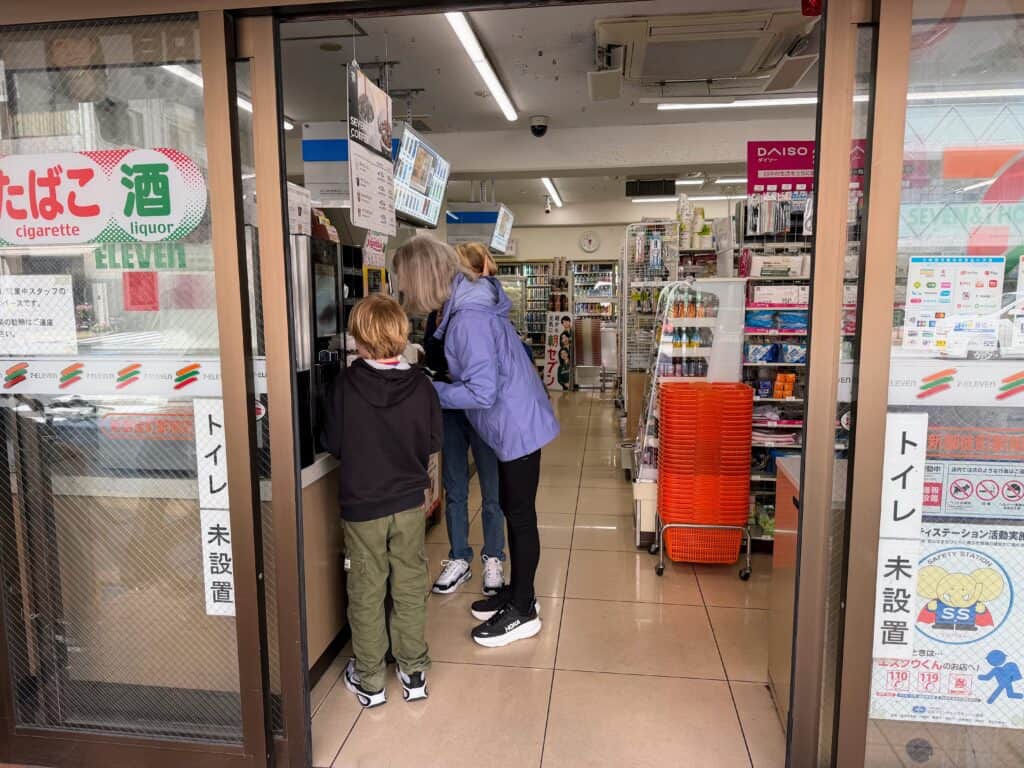 Woman in purple rain coat and two boys look at something on the counter in 7-11 in Japan.