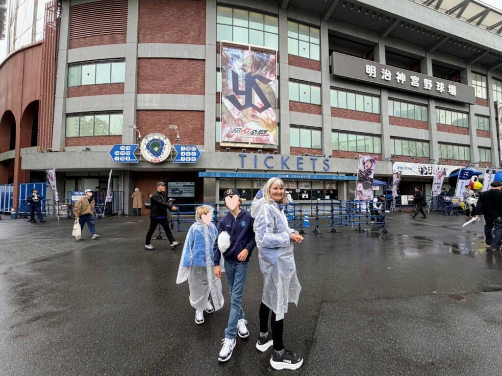 Grandmother and grandsons wear rain ponchos in front of Tokyo Meiji Jingu baseball stadium on a Yakult Swallows game day.