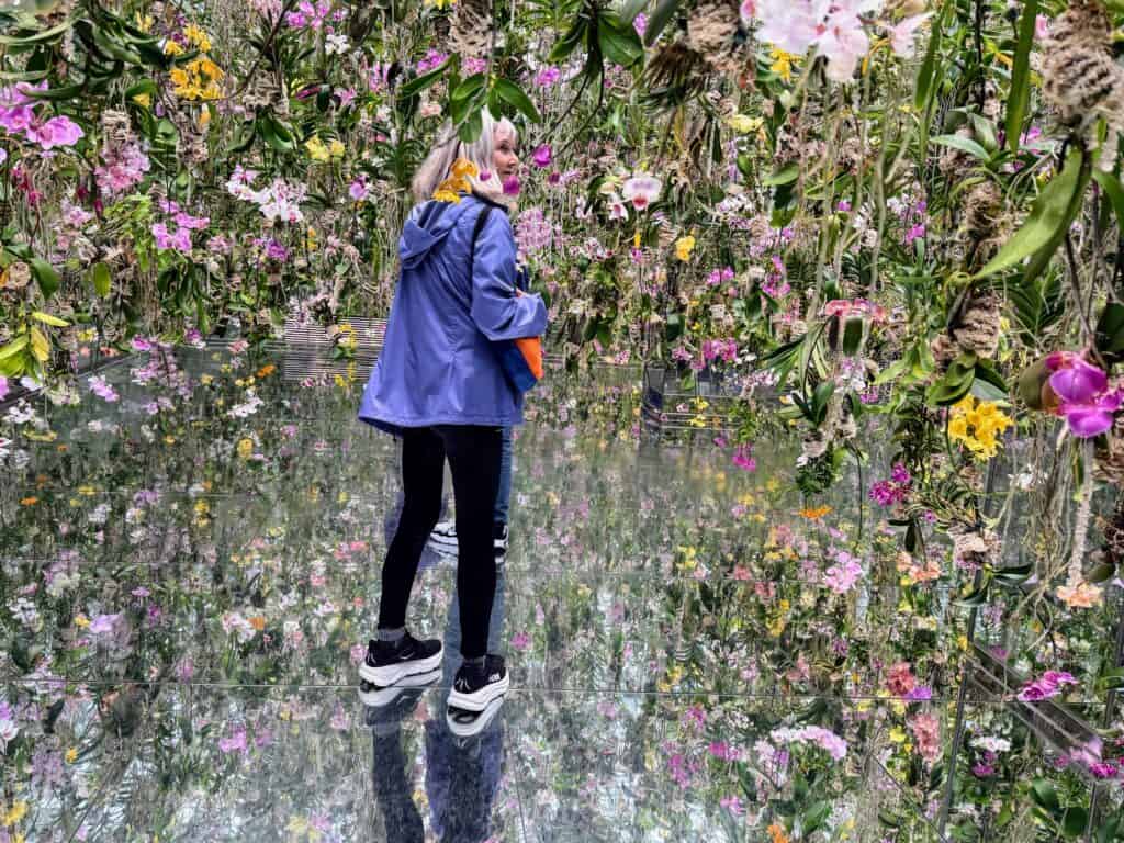 Woman in purple coat stands among orchids that are hanging from the ceiling in an exhibit in Tokyo's Teamlab Planets. The floor is a mirror.