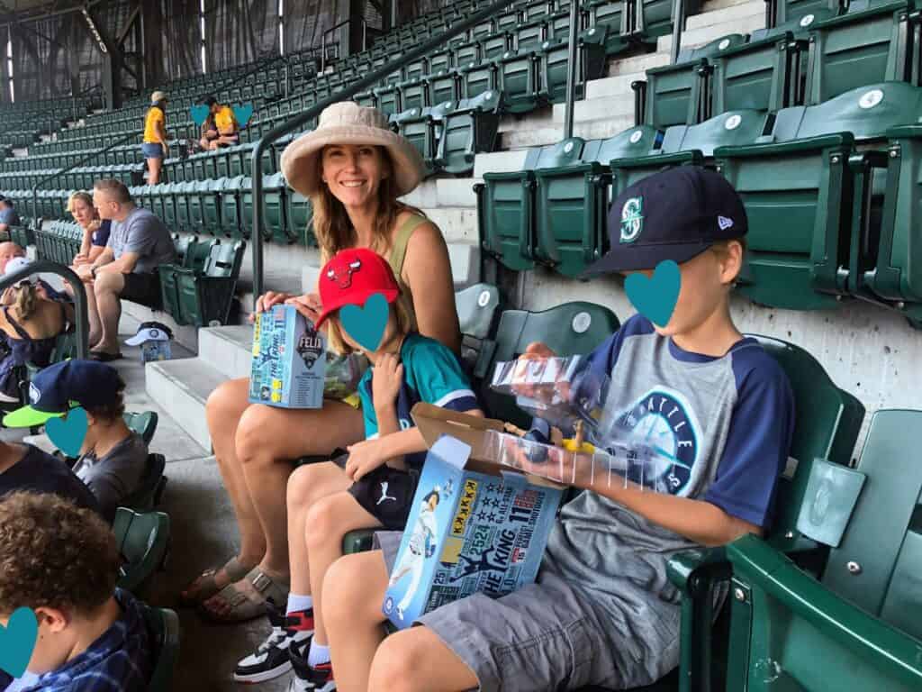 Mom in hat sits in Seattle Mariners baseball stadium stands with two children. They are looking at bobbleheads.