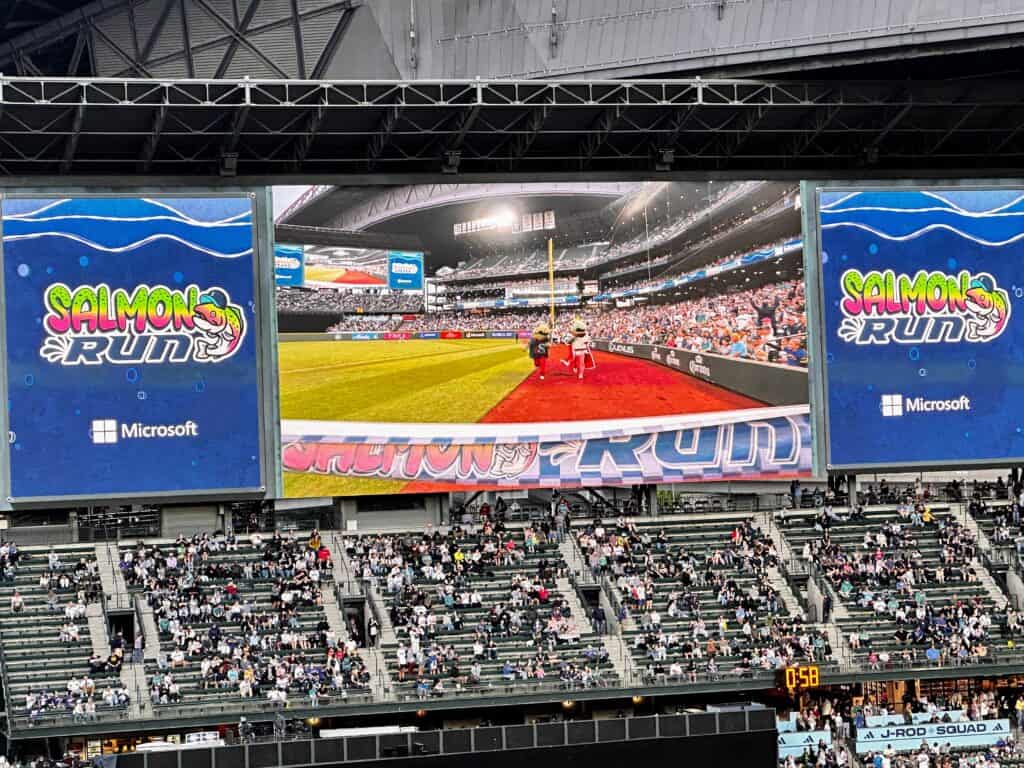 Close up of digital scoreboard at Seattle Mariners baseball stadium. It shows the Salmon Run race on the field. People in stands below the scoreboard.