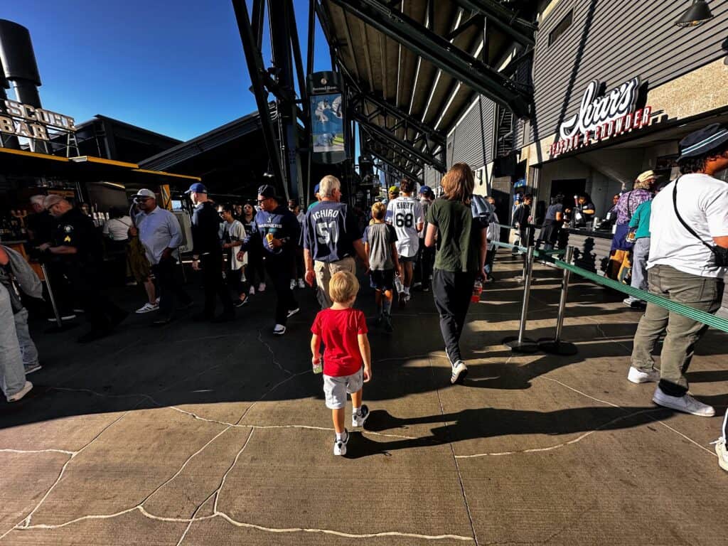 Boy follows crowd in outdoor area of upper deck of Seattle Mariners baseball stadium. Ivars seafood concessions on right and bar on left.