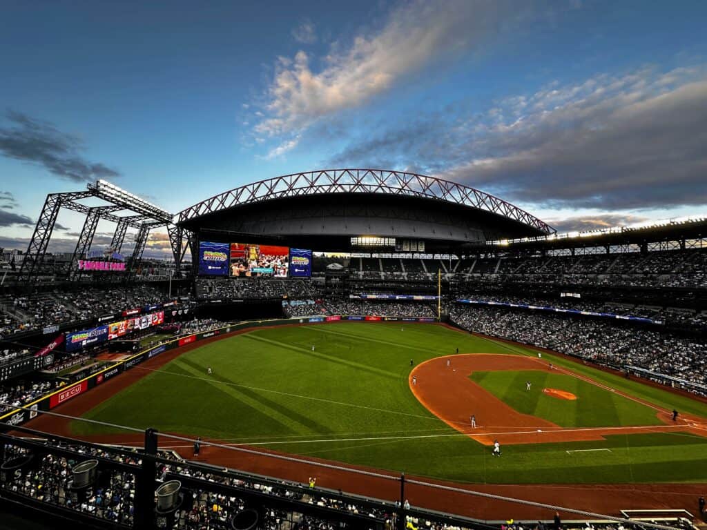 Seattle Mariners baseball stadium at dusk in the summer. Players are on the field and the stands are full of fans.