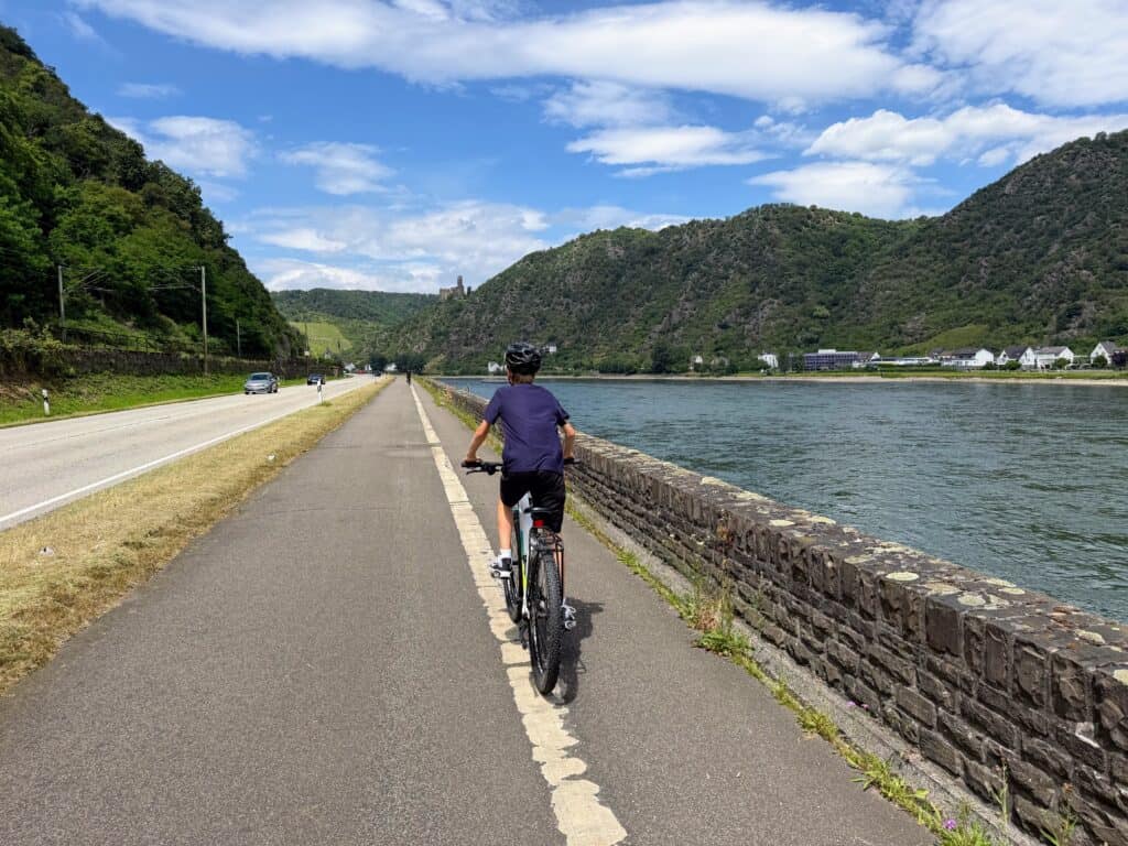 Boy in dark blue shirt rides on a cycling path along the Rhine River in Germany. Road and car to the left of the path. River to the right and castle on a hill ahead in the distance.
