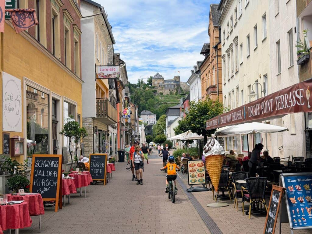 Boys cycle down pedestrian area of the center of St. Goar in Germany. Ice cream cone marks an ice cream shop on the right and other eateries are open with outdoor seating on either side. Castle in the distance on a hill.