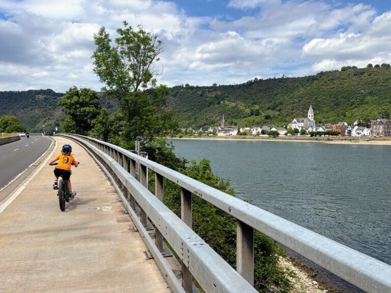 Young boy in orange jersey rides his bike along a cycling path with the Rhine River on the right. A town with a church steeple towering above and green hillsides are on the opposite side of the river. Road to the left of bike path.