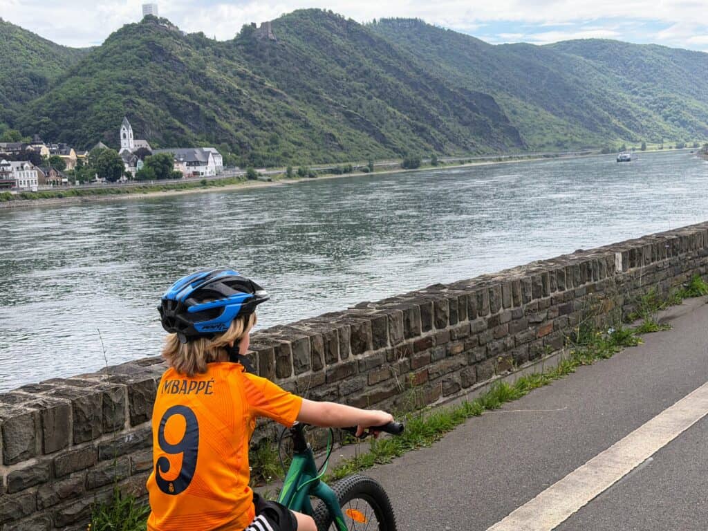 Boy in orange shirt and blue helmet cycles on a paved path next to the Rhine River in Germany. The opposite side of the river has green hillsides and a small village with a church and its large white steeple.