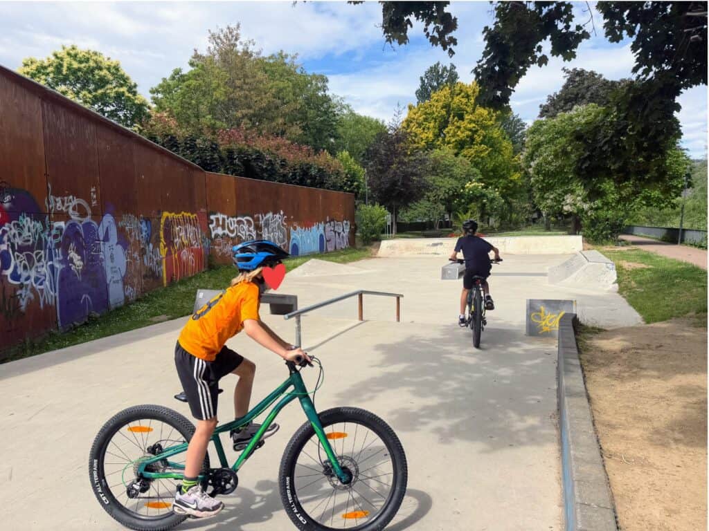 Two boys ride bicycles at a skate park in Boppard, Germany. Graffiti on metal wall on left and leafy trees surrounding the park.