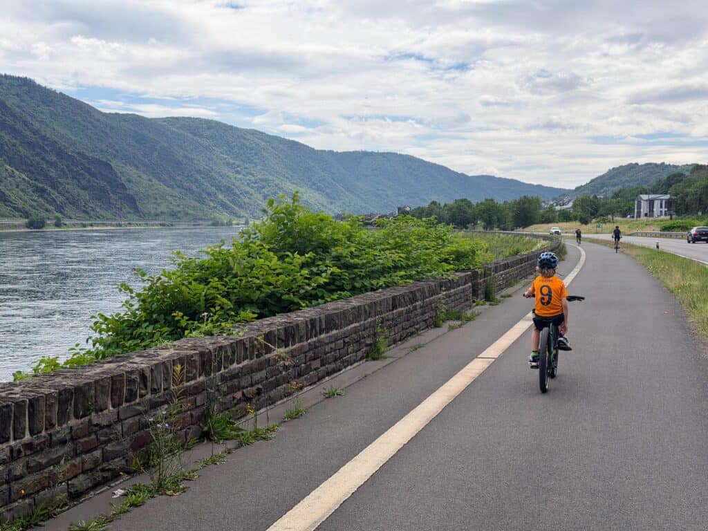 Boy in orange shirt rides his bicycle one-handed along a cycling path on the Rhine River (on the left). Others cycle ahead of him. The road with a couple of cars is to the right of the path. Green hills on the other side of the river.