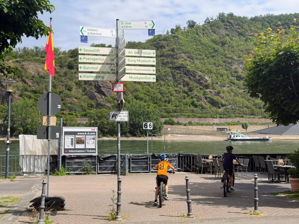 Boys ride on cobblestones toward the river (Rhine). White road signs are behind them, and some are pointing toward the bicycle paths.