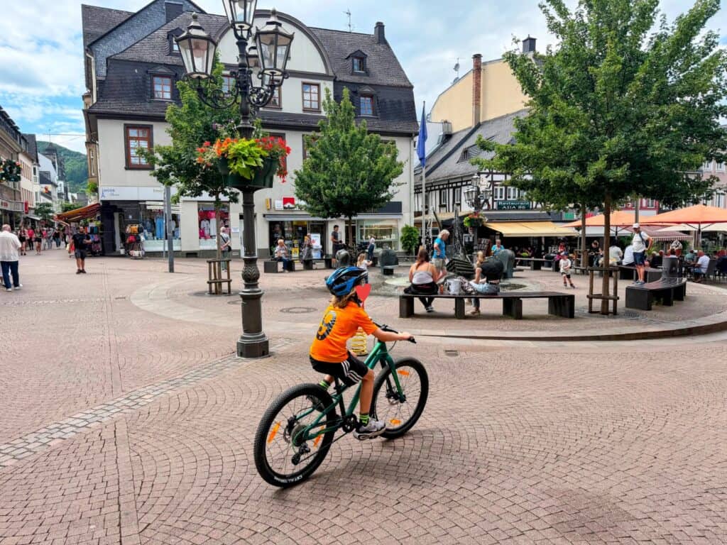 Boy in orange shirt rides his bicycle on a cobblestone square past people sitting on benches and playing in a small fountain. A few trees in the square, and German buildings on the opposite side of the square.