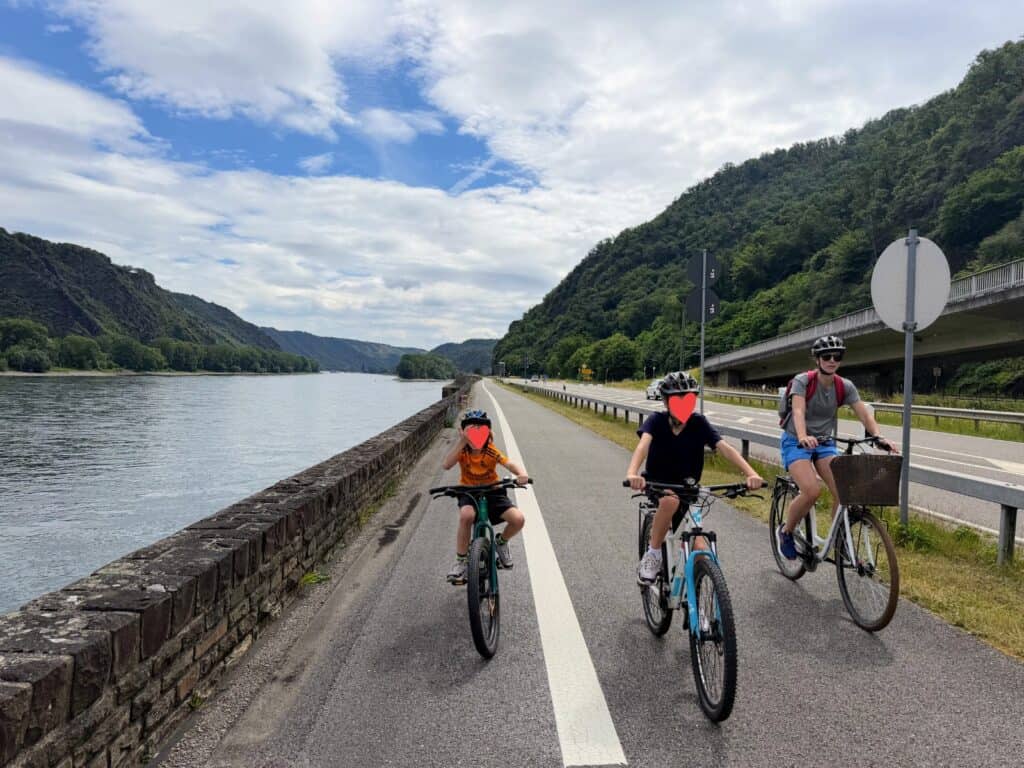Two boys and their mother cycle toward the camera on a paved path along the Rhine River in Germany. The river is to the left and a paved road is on the right.