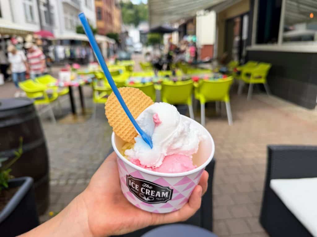 Child's hand holds a cup of lemon and strawberry gelato in front of outdoor seating at the shop in the pedestrian area of St. Goar in Germany.