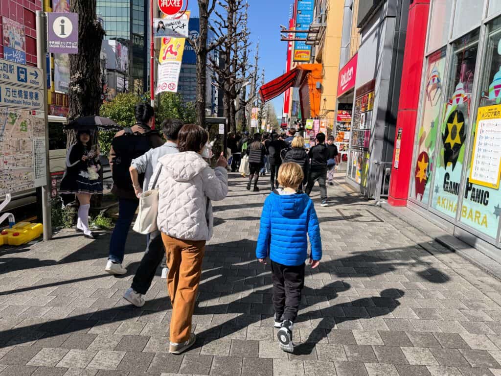 Guide and boy walk down sidewalk in Akihabara, Tokyo. There are colorful signs on sides of sidewalk and other people walking ahead of them. It's a sunny day.