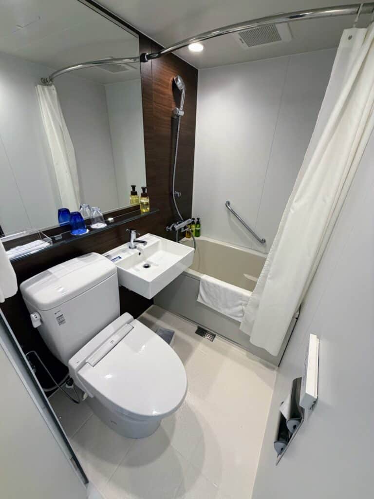 Bathroom at HOTEL MYSTAYS Fuji Onsen Resort. Toilet with sink, mirror, and bath/shower. Toiletries in large containers above sink and in shower.