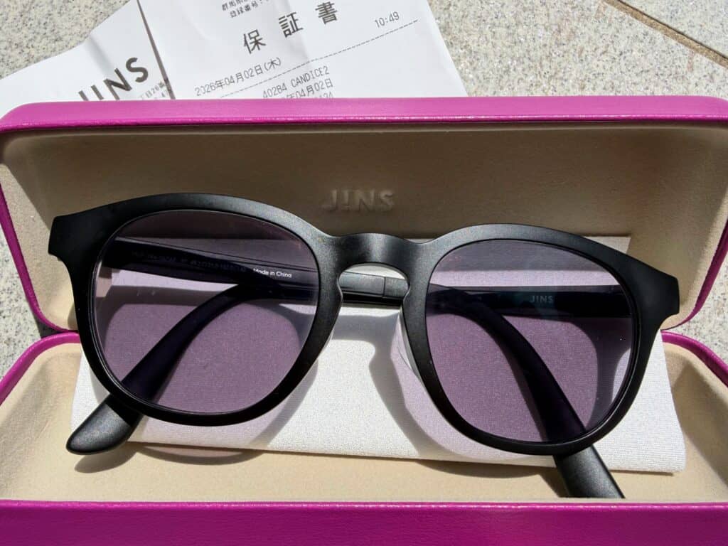 Hot pink hard eyeglasses case with a pair of black rimmed glasses inside. The case is sitting on a stone surface and there is a receipt from JINS underneath.