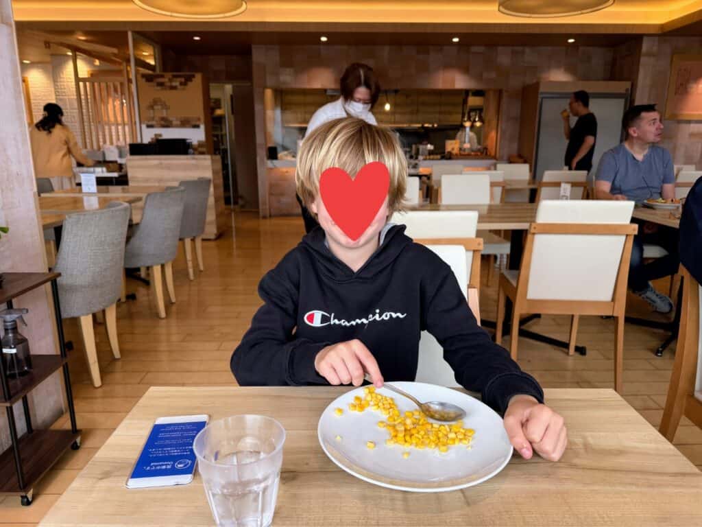 Boy sits at wooden table in breakfast area at HOTEL MYSTAYS Fuji Onsen Resort. He has a white plate with corn and a glass of water. Other people dine at tables behind him.
