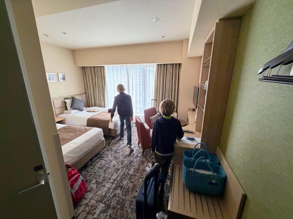 Boys walk into room at HOTEL MYSTAYS Fuji Onsen Resort. They are looking toward window which is covered with a shade. Two beds, two chairs, shelving unit on right.