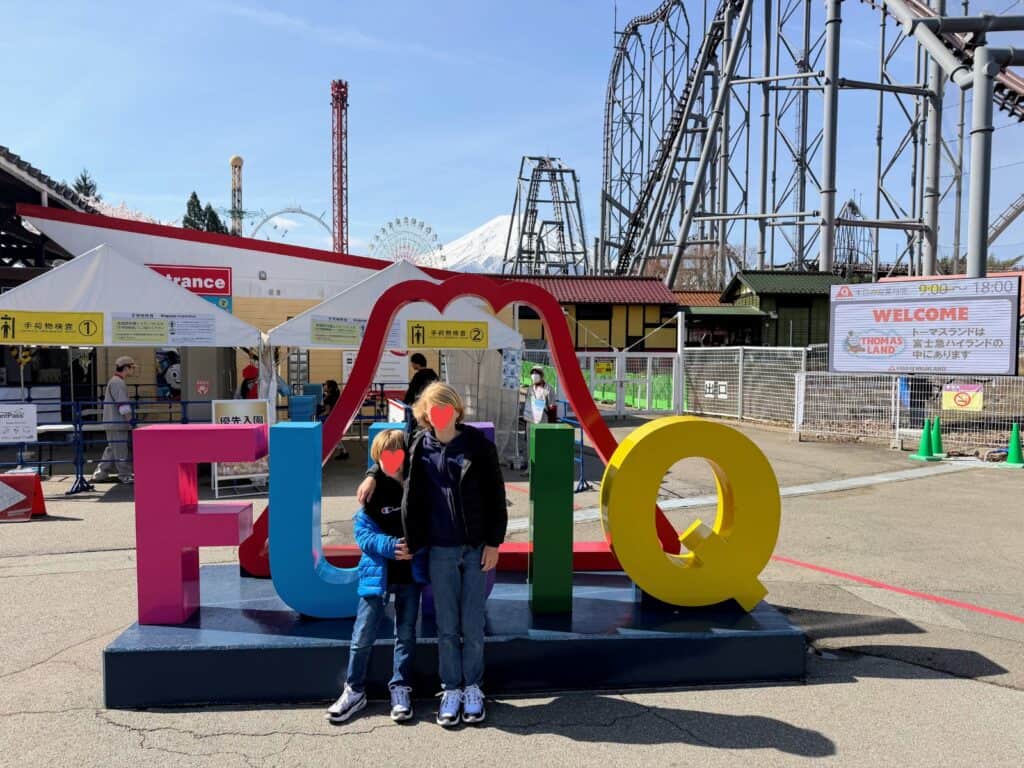 Two boys stand in front of colorful sign for Fuji-Q. Behind are buildings, entrance tents, large rollercoasters, and Mt. Fuji at Fuji-Q Highland theme park in Japan.