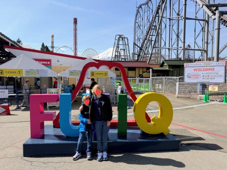 Two boys stand in front of colorful sign for Fuji-Q. Behind are buildings, entrance tents, large rollercoasters, and Mt. Fuji at Fuji-Q Highland theme park in Japan.