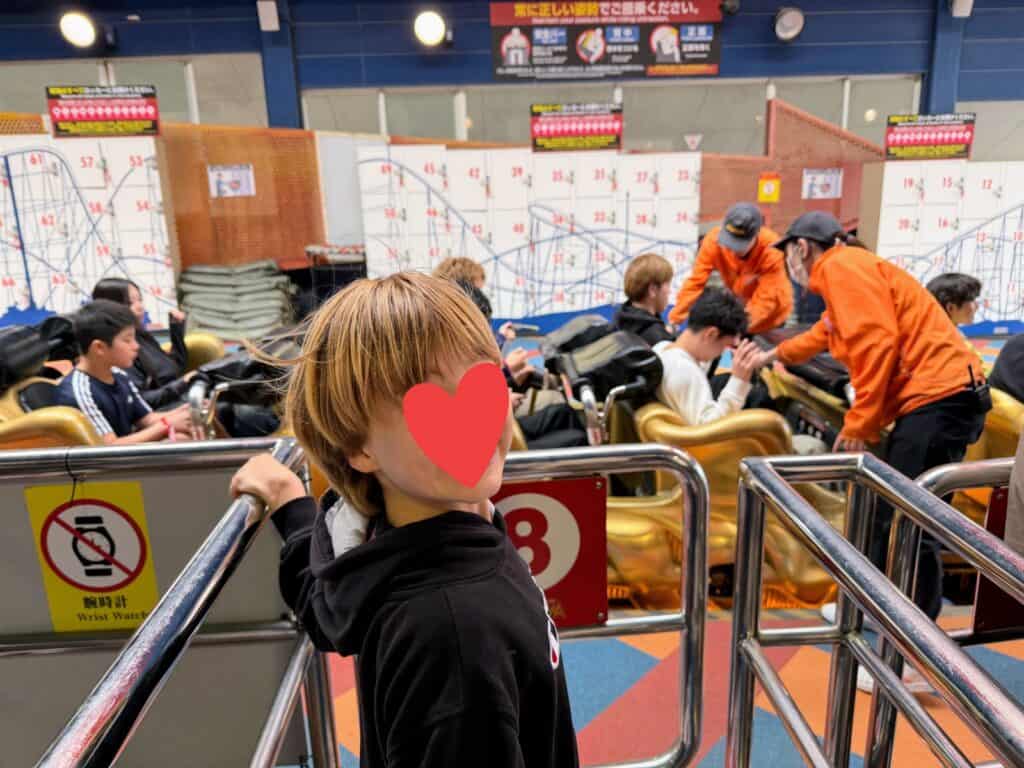 Boy stands at front of line, ready to get on the Fujiyama rollercoaster at Fuji-Q Highland theme park in Japan. Others are in the coaster getting their seatbelts checked by staff.