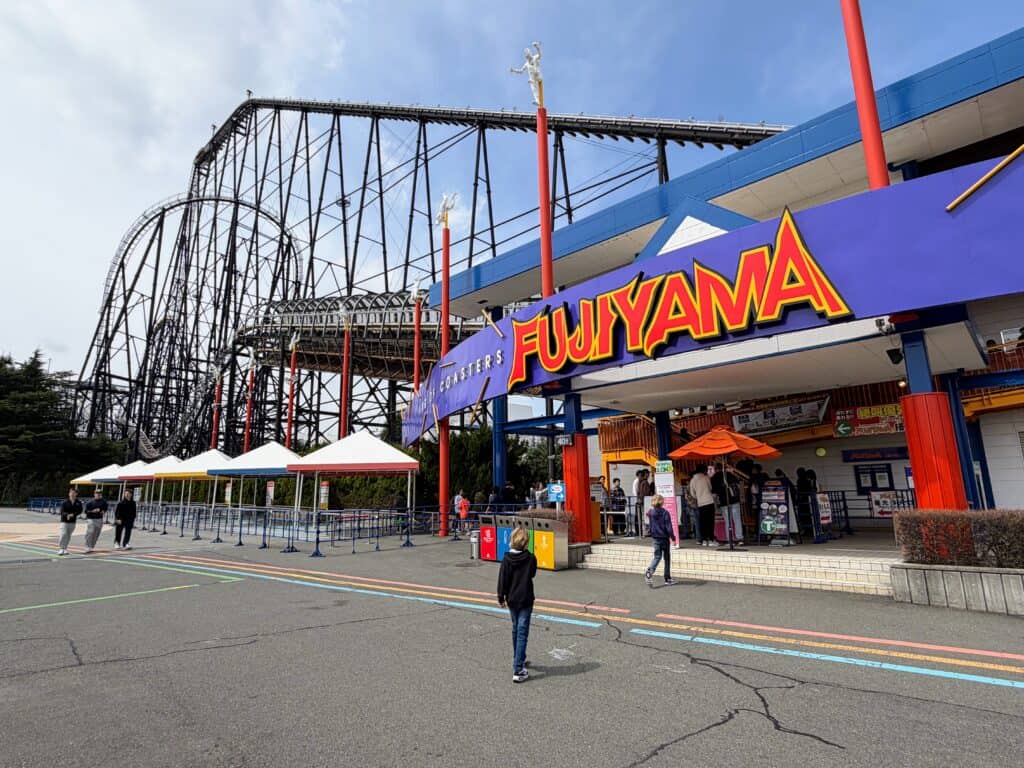 Boys walk toward the entrance to the Fujiyama rollercoaster at Fuji-Q Highland theme park in Japan. A few others wait in line and the rollercoaster looms behind the building entrance. Paved area out front and a few other people are walking by.