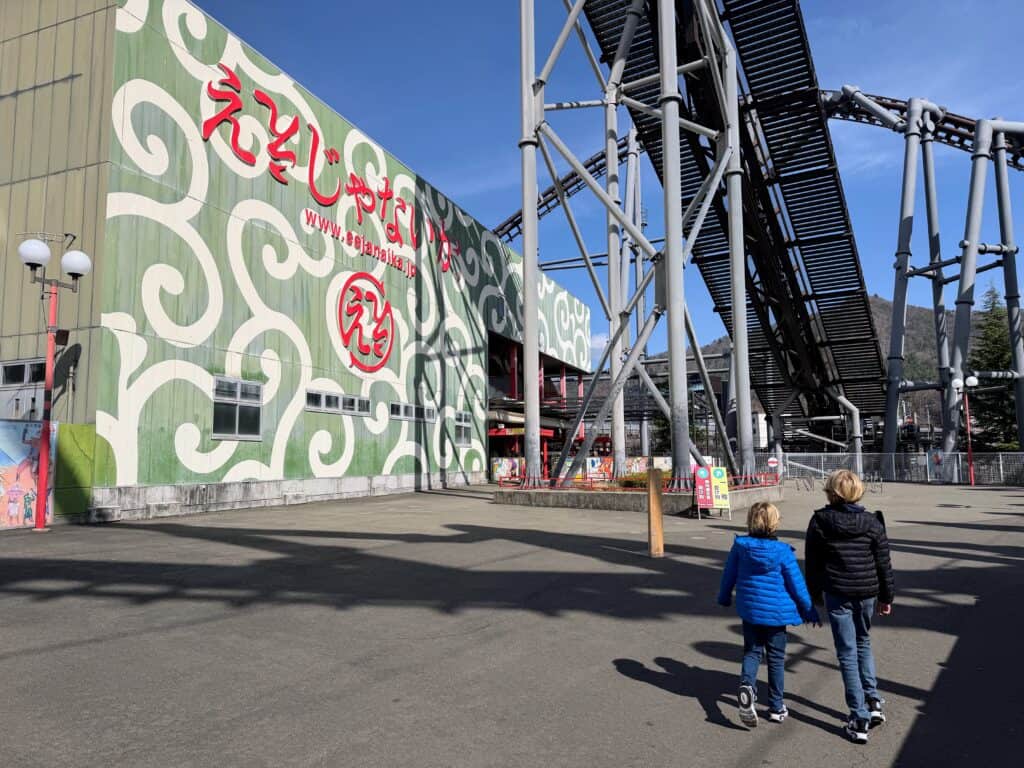 Boys in pants and coats walk toward the Eejanaika rollercoaster at Fuji-Q Highland theme park in Japan. The rollercoaster track rises sharply above them. There is a large green building with squiggly white lines to the left.