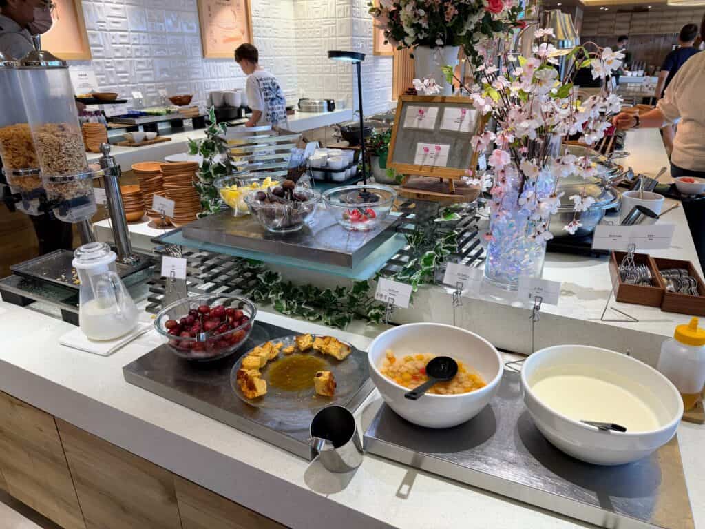 Breakfast buffet at HOTEL MYSTAYS Fuji Onsen Resort. Fresh fruit in bowls, cereal, silverware, milk, yogurt. Flowers on the display.