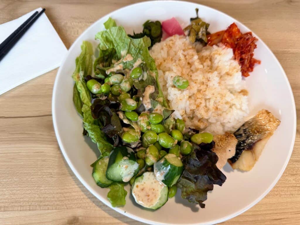 Breakfast plate at HOTEL MYSTAYS Fuji Onsen Resort. White plate with salad, soybeans, rice, kimchee and fish on a wooden table. Chopsticks to side.