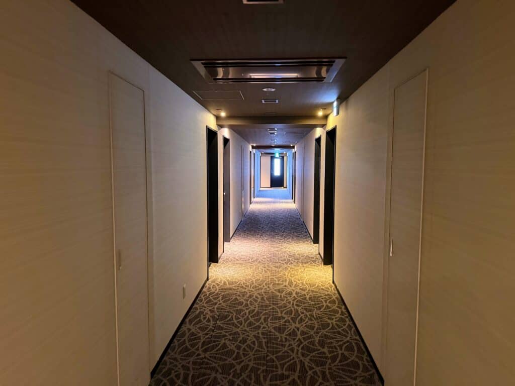 Long hallway at HOTEL MYSTAYS Fuji Onsen Resort. Cream colored walls and brown patterned carpet.