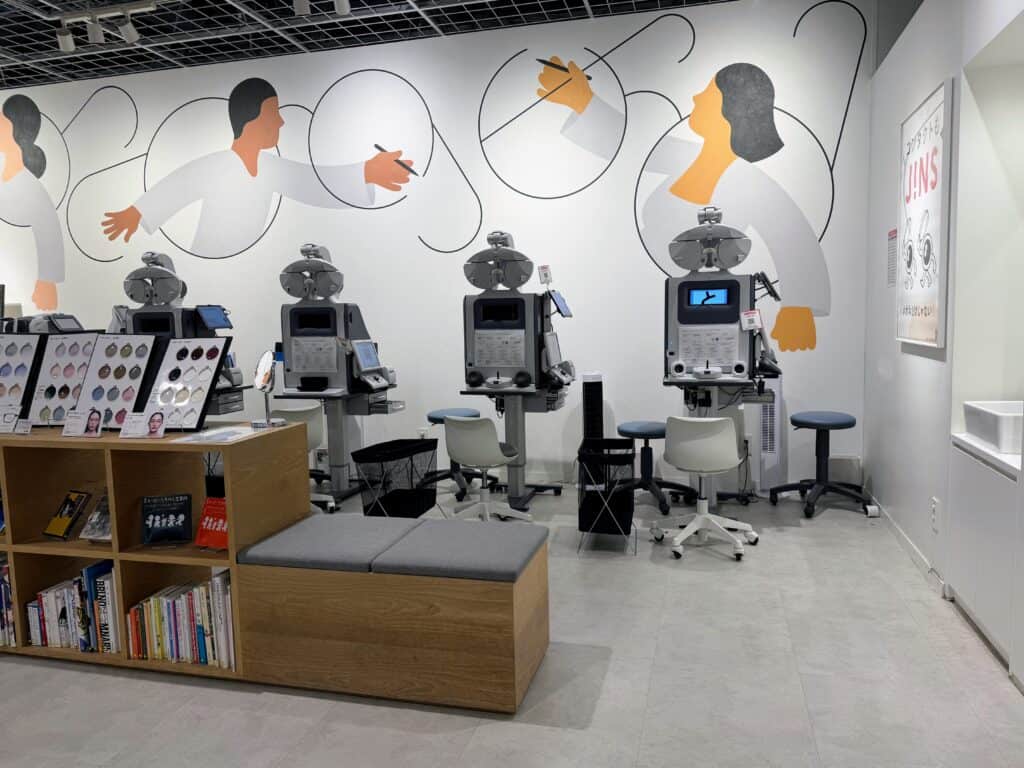 Four grey machines lined up against a wall. The machines are used for measuring prescriptions for eyewear. In front of the machines is a bench seat and to the left, small bookshelves and displays of tinted lenses for eyeglasses. The wall behind the grey machines is white with cartoon people "drawing" eyeglass frames. This is inside a JINS glasses store in Tokyo, Japan.