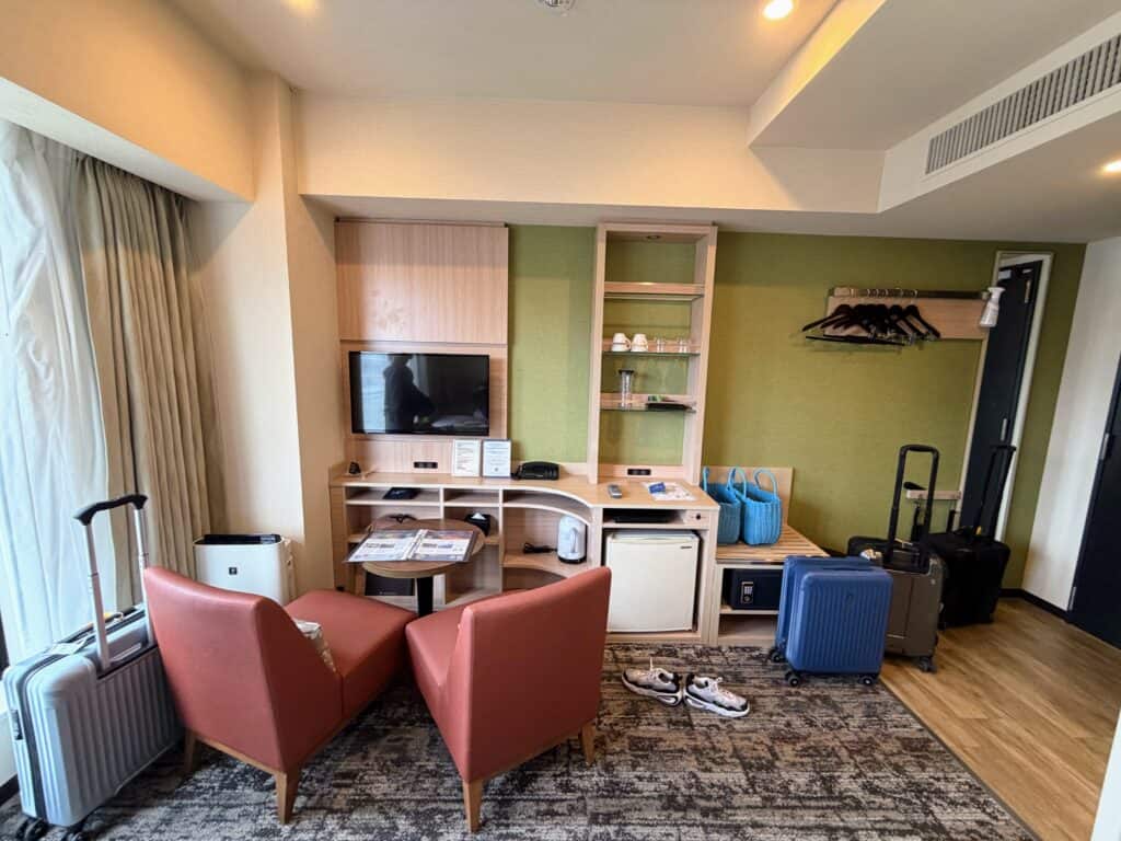 Living area in room at HOTEL MYSTAYS Fuji Onsen Resort. Two red chairs at a small table, suitcases unopened on floor, shelving unit has fridge, tv, kettle, cups, etc. Also hangers and spa bags.