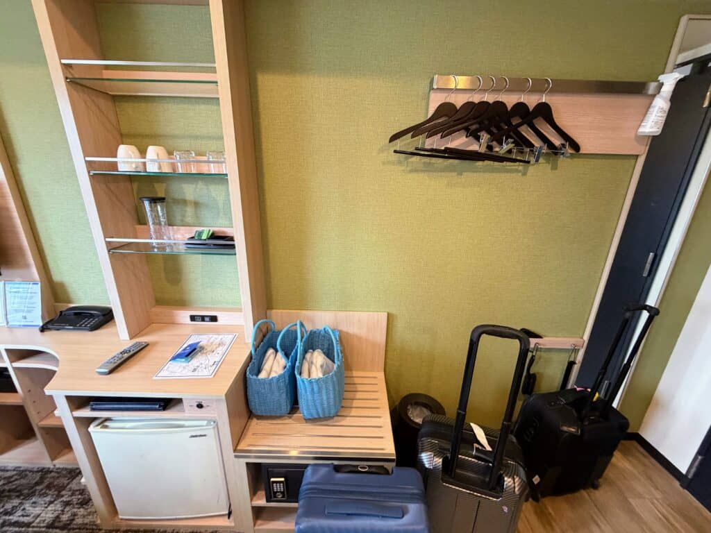 Storage area and dining area (with fridge and cups) at HOTEL MYSTAYS Fuji Onsen Resort. Hangers on rack on wall, blue baskets on a shelf hold spa towels. Suitcases on floor. Mini fridge on left. Cups on shelves above fridge. In a room at HOTEL MYSTAYS Fuji Onsen Resort.
