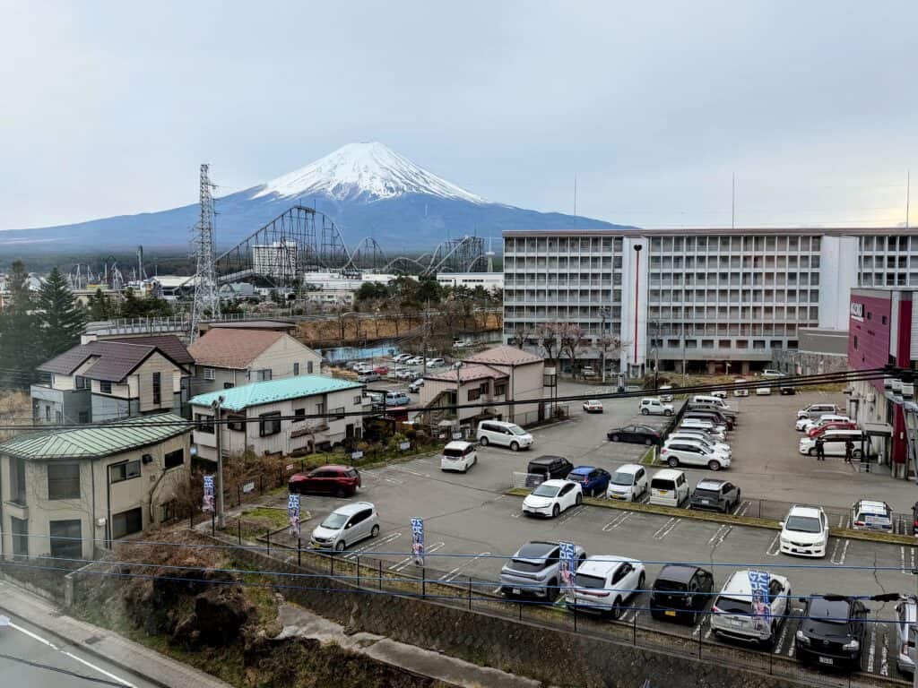 Snow-capped Mt. Fuji. In front of it are roller coasters of Fuji-Q Highland theme park and a couple of multi-story buildings and a parking lot. Other smaller buildings to left of parking lot.