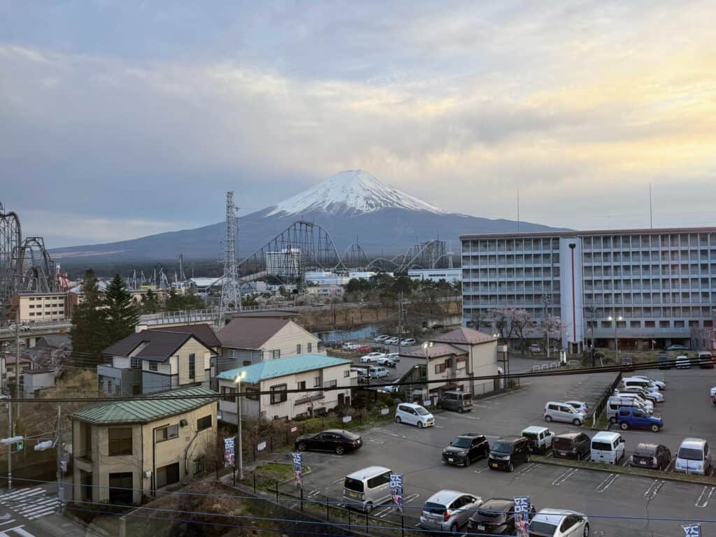 View of snow-capped Mt. Fuji, a theme park with roller coasters, buildings, and a parking lot.