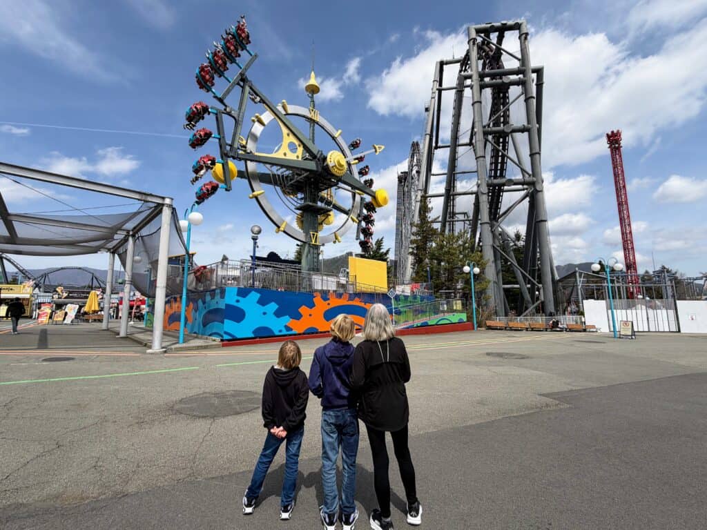 Grandmother and grandsons watch people on a ride at a theme park in Japan (Fuji-Q Highland). Large rollercoaster to the right of the ride they are watching.