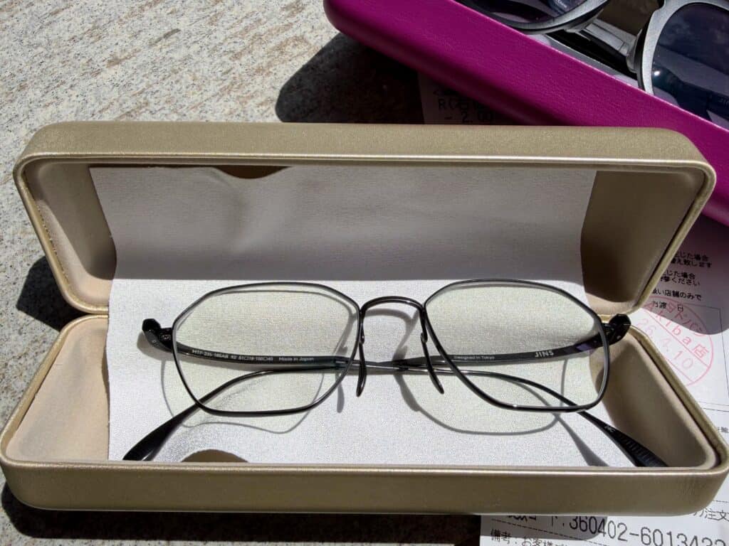 Pair of wire-rimmed eye glasses in an opened gold case. In corner is a pair of black glasses in a hot pink case.