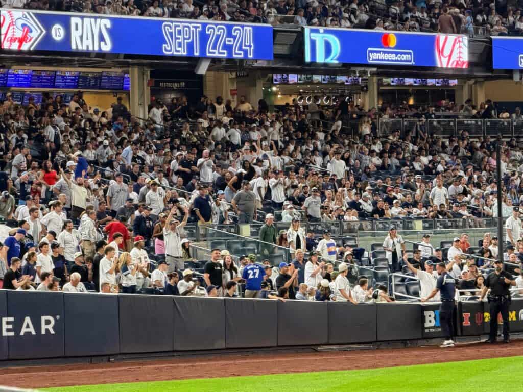 First baseline stands at Yankee Stadium. Fans crowd the first row asking for a ball from players or the ball boy.