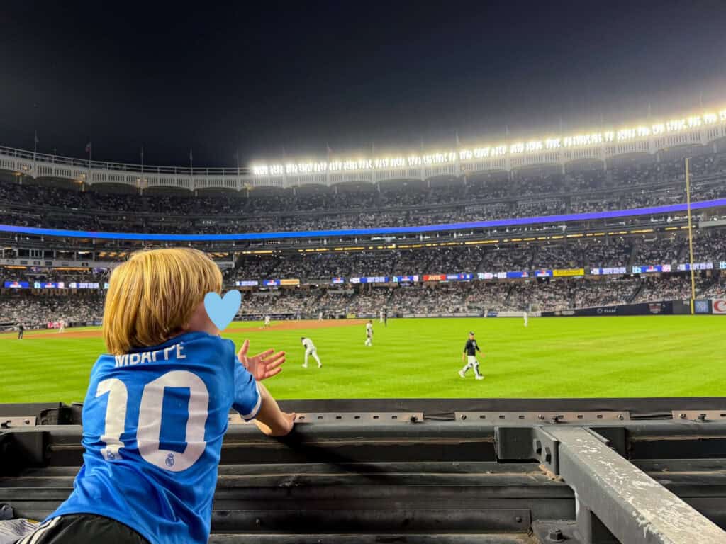Boy in Mbappe soccer jersey leans over an outfield wall in the stands at Yankee Stadium while players warm up between innings. It's evening. He holds his hand out asking for a ball from a player.