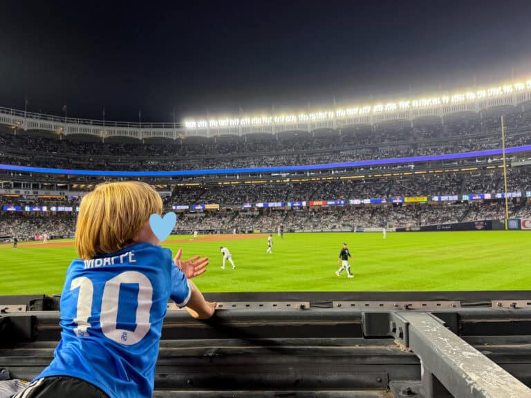 Boy in Mbappe soccer jersey leans over an outfield wall in the stands at Yankee Stadium while players warm up between innings. It's evening. He holds his hand out asking for a ball from a player.