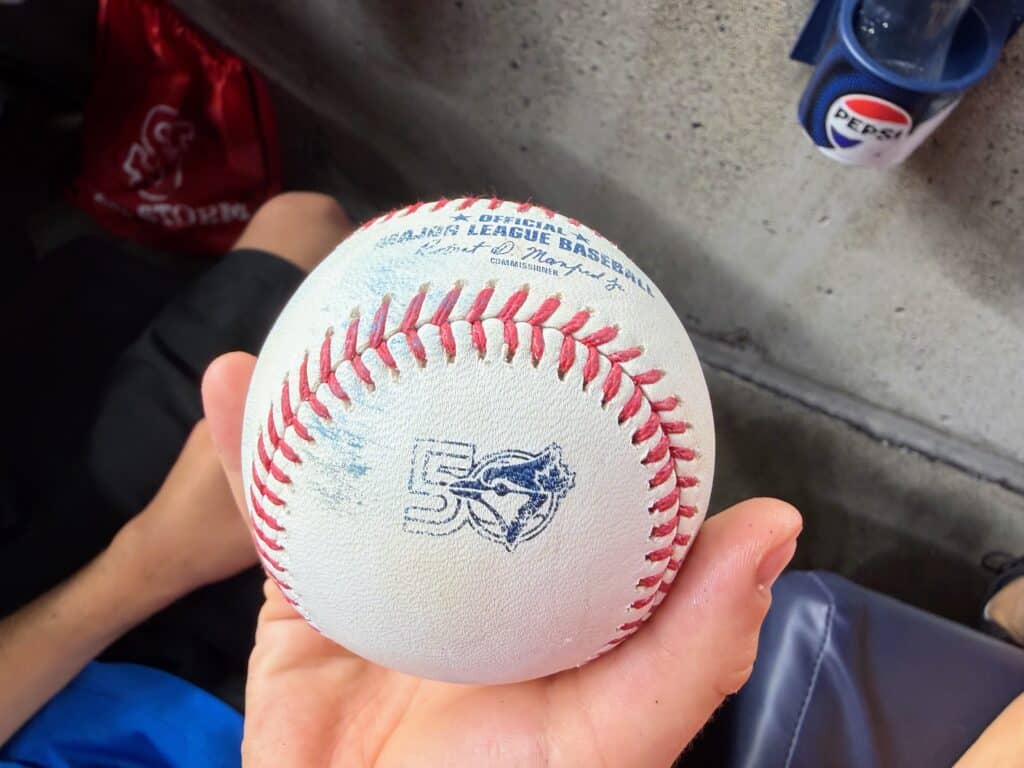 Child's hand holds a MLB baseball with a 50 and Toronto Bluejays logo stamped on it.