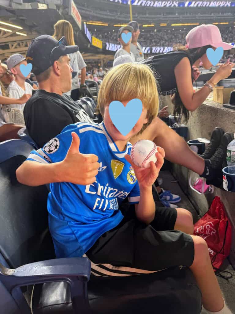 Boy in blue shirt sits in stands at Yankee Stadium. He's holding a baseball and giving a thumbs up.
