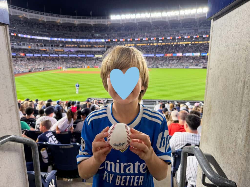 Boy stands with a baseball in his hands and Yankee Stadium stands behind him. He's in the outfield stands. It's night and the stadium is lit up and packed with fans.