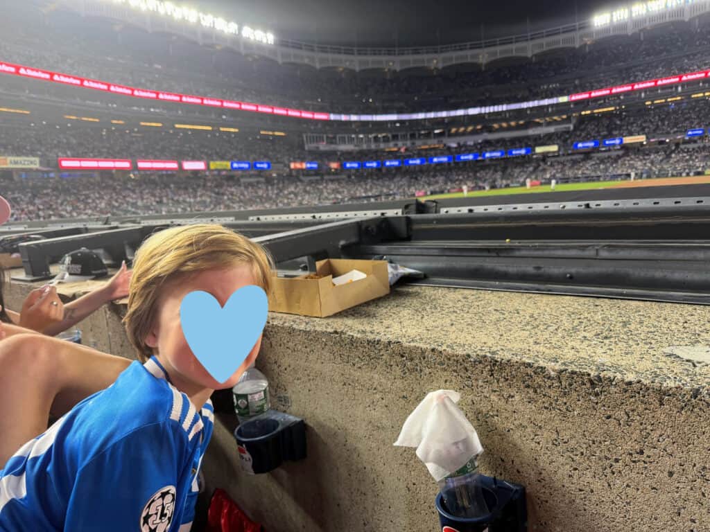 Boy in blue shirt sitting behind concrete wall at Yankee Stadium at night. Stands are full and lit up with stadium lights.