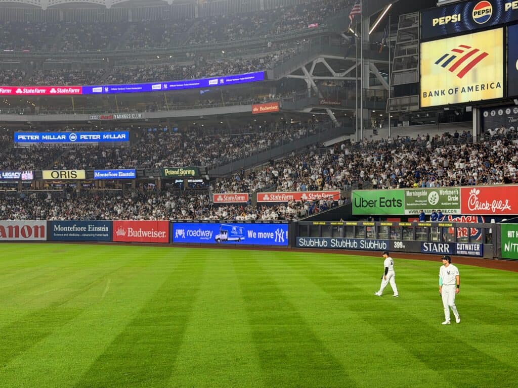 Outfield stands packed with fans look out onto the field. There are two NY Yankees players standing in the outfield.